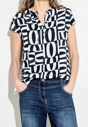 Kvinde iført en kortærmet sort og hvid mønstret bluse, stoppet ned i mørkeblå denimjeans med hænderne i lommerne.