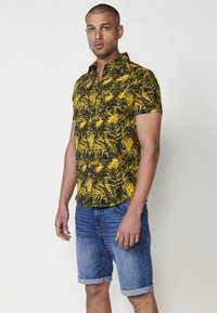 Cămașă cu mânecă scurtă, cu un model de frunze de palmier alb-negru și galben, având guler și nasturi, asortată cu pantaloni scurți din denim.