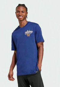 Jaunas vyras su nupintomis plaukų kasomis, vilkintis mėlyną Adidas marškinėlį su užrašu "nuo 1949", besišypsantis, atsipalaidavęs rankomis, šviesiame fone.