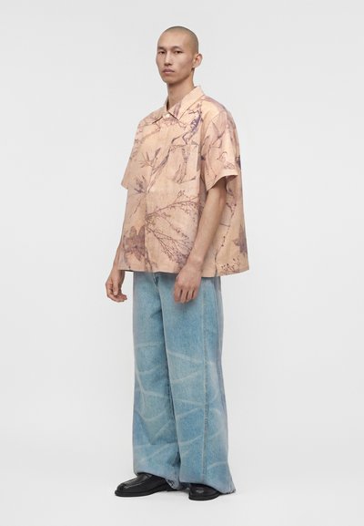 Άτομο στέκεται πλαγίως φορώντας φαρδιά μπεζ κοντομάνικη μπλούζα με λουλούδια, φαρδιά ανοιχτό μπλε τζιν και μαύρα παπούτσια σε απλό φόντο.