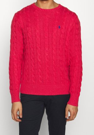 Mies, jolla on päällään kirkkaanpunainen kaapelineulottu neule, jossa sininen logo rinnassa ja mustat housut, seisomassa valkoista taustaa vasten.