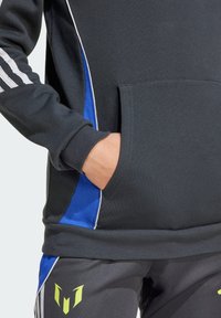 Sweatshirt noir avec des panneaux latéraux bleus, des rayures blanches et une poche frontale. Associé à un pantalon sombre arborant un logo jaune sur la cuisse.