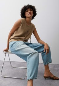 Woman with curly hair sits on a metal chair wearing a sleeveless brown buttoned vest, light blue jeans, and brown heeled sandals.