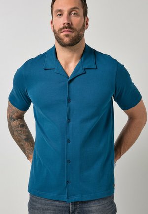 Uomo con barba e braccio tatuato che indossa una camicia verde acqua a maniche corte con bottoni e sta in piedi con le mani dietro la schiena.