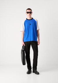 Blue short-sleeve shirt with white accents and a black collar, paired with black trousers and sunglasses. Holds a black bag. Smooth texture.