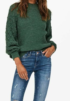 Femme portant un pull en maille texturée vert foncé avec des manches ballon et un jean skinny bleu déchiré.