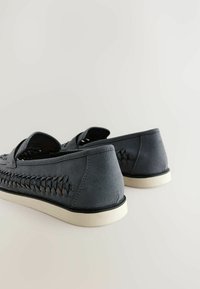 Zapatos de ante gris con detalles trenzados en los lados, suelas blancas planas y bordes redondeados. Diseño de parte superior abierta y ajuste relajado.
