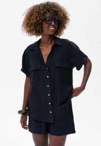 Czarna lniana koszula z guzikami, z dwoma przednimi kieszeniami, krótkimi rękawami i luźnym krojem. Połączona z dopasowanymi szortami; drewniane guziki i detale.