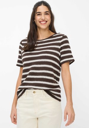 Vrouw lacht en draagt een bruin-wit gestreept gebreid topje met korte mouwen en lichtgekleurde broek met hoge taille tegen een effen achtergrond.