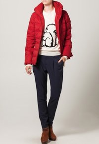 Rød pufferjakke med hætte, båret over en beige sweater med et egerndesign. Kombineret med marineblå bukser og brune ankelstøvler.