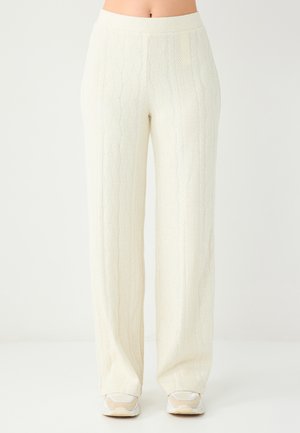 Spodnie o szerokich nogawkach w kolorze ecru z teksturowanym wzorem warkocza. Posiadają średni stan i delikatny połysk. Połączone z jasnymi sneakersami.