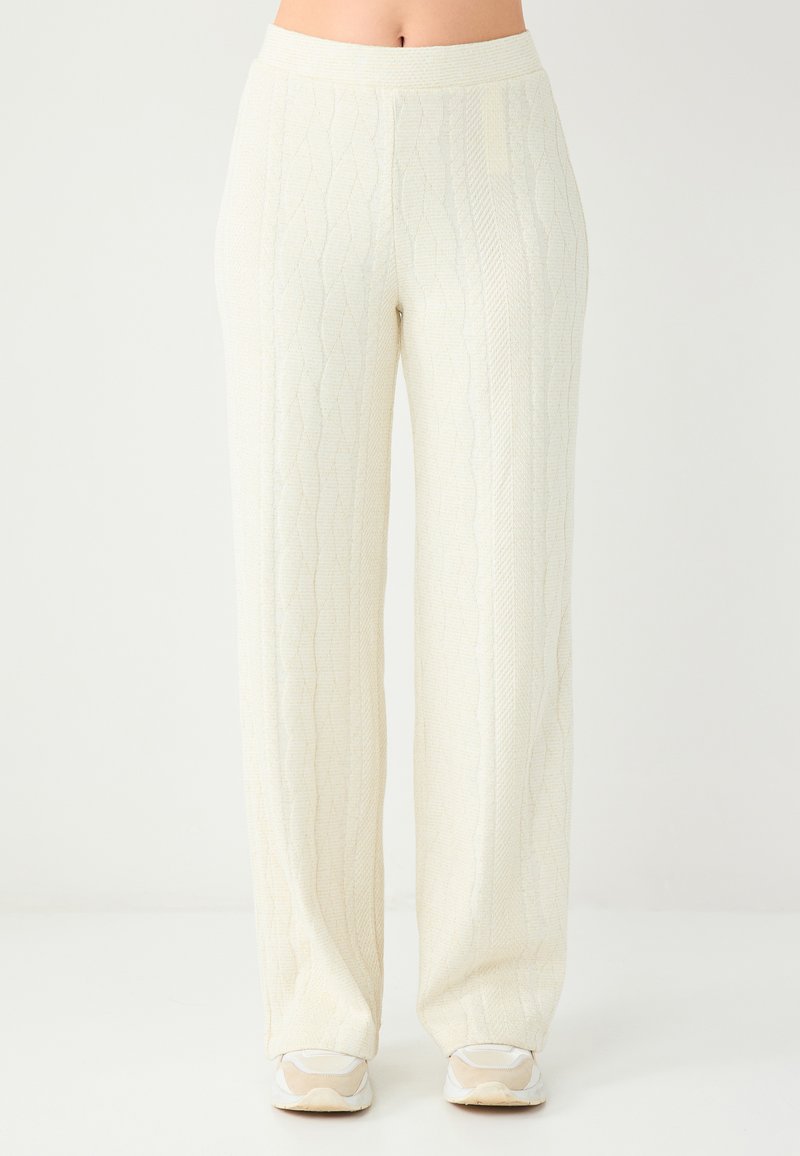 Pantalon à jambes larges de couleur blanc cassé avec un motif texturé en tricot torsadé. Il présente une taille mi-haute et un léger éclat. Associé à des baskets claires.