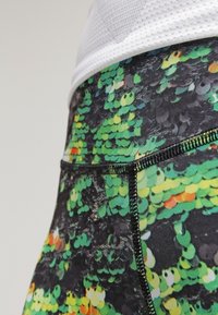 Funkční legíny s černým základem a živým vzorem připomínajícím flitry v zelené a žluté barvě. Mají černé švy a hladkou strukturu.