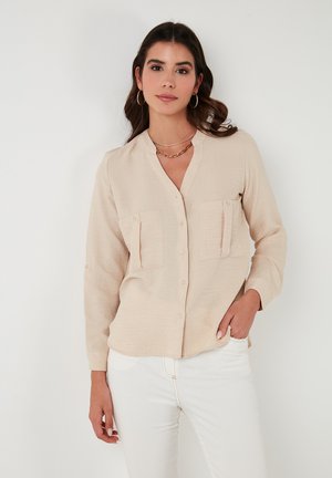 Light beige long-sleeve shirt with a V-neck, two chest pockets, and button closures. Fabric has a textured appearance. Worn with white pants.