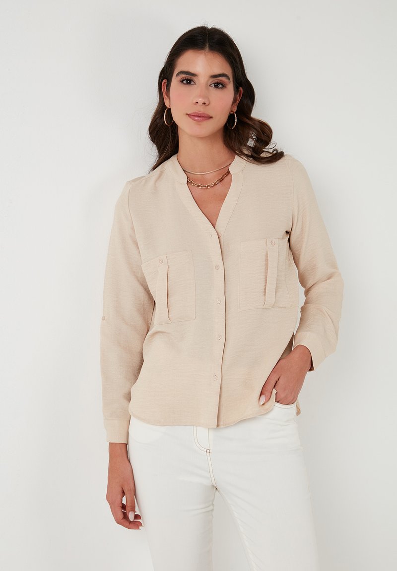 Light beige long-sleeve shirt with a V-neck, two chest pockets, and button closures. Fabric has a textured appearance. Worn with white pants.
