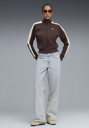 Braune Trainingsjacke mit Reißverschluss und cremefarbenen Streifen, kombiniert mit hellblauen Jeans mit weitem Bein und beigen Schuhen mit braunen Sohlen. Accessoires umfassen eine Sonnenbrille.