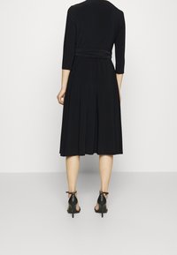Černé midi šaty se třičtvrtinovými rukávy, plisovanou sukní a zavázaným pasem. Nosené s černými sandály na podpatku. Hladká textura, minimalistický design.