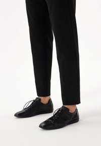 Persoană purtând pantaloni negri conici și pantofi casual negri din piele cu șireturi, stând pe un podea albă.