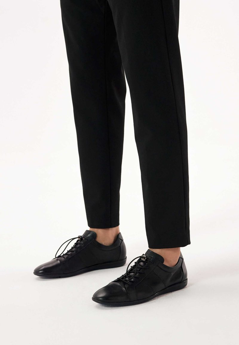 Persoană purtând pantaloni negri conici și pantofi casual negri din piele cu șireturi, stând pe un podea albă.