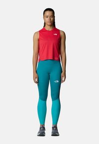Top sin mangas rojo, leggings verde azulado, ambos ajustados. El top tiene un dobladillo redondeado; los leggings tienen un logotipo en el costado. Se lleva con zapatillas de deporte grises.