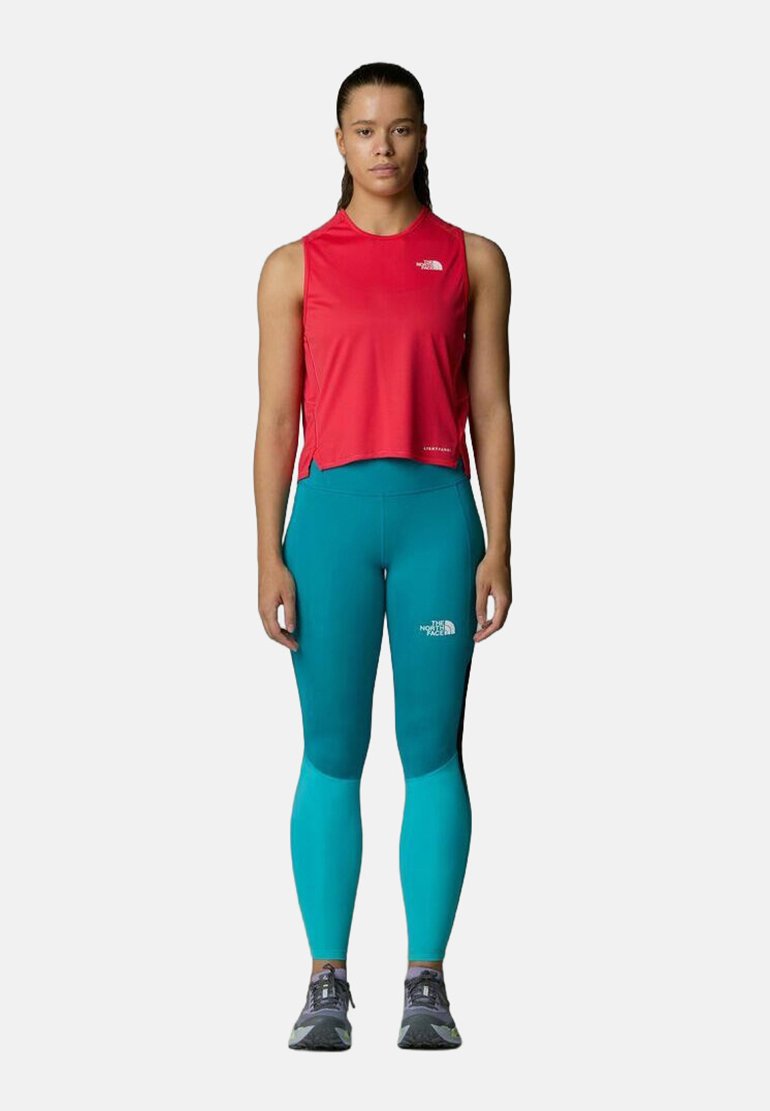 Top sin mangas rojo, leggings verde azulado, ambos ajustados. El top tiene un dobladillo redondeado; los leggings tienen un logotipo en el costado. Se lleva con zapatillas de deporte grises.