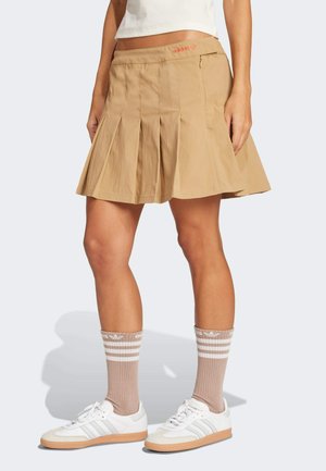 Pleated tan skirt featuring a smooth texture, zip pocket, and an embroidered red logo. Paired with white sneakers and striped brown socks.