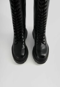 Des bottes hauteur genou en cuir noir avec une semelle épaisse, un laçage prononcé sur le devant et une finition brillante.