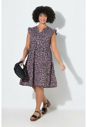 Kobieta idąca w fioletowej sukience do kolan w panterkę, trzymająca czarną torebkę, nosząca czarne sandały na platformie.
