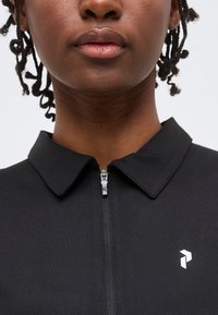 Κοντινό πλάνο ενός ατόμου που φοράει μαύρο πουκάμισο με φερμουάρ, γιακά και μικρό λευκό λογότυπο στο στήθος.