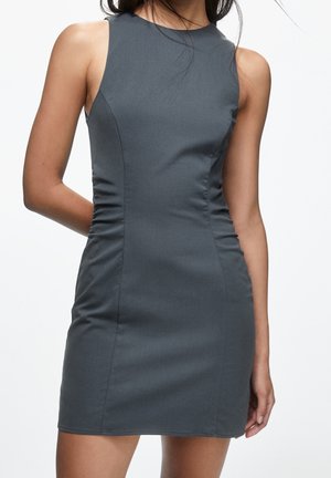 Ærmeløs tætsiddende mørkegrå kjole på en kvinde, med høj halsudskæring og rynkede sidesømme, hvor kvindens hånd hviler bag på ryggen.