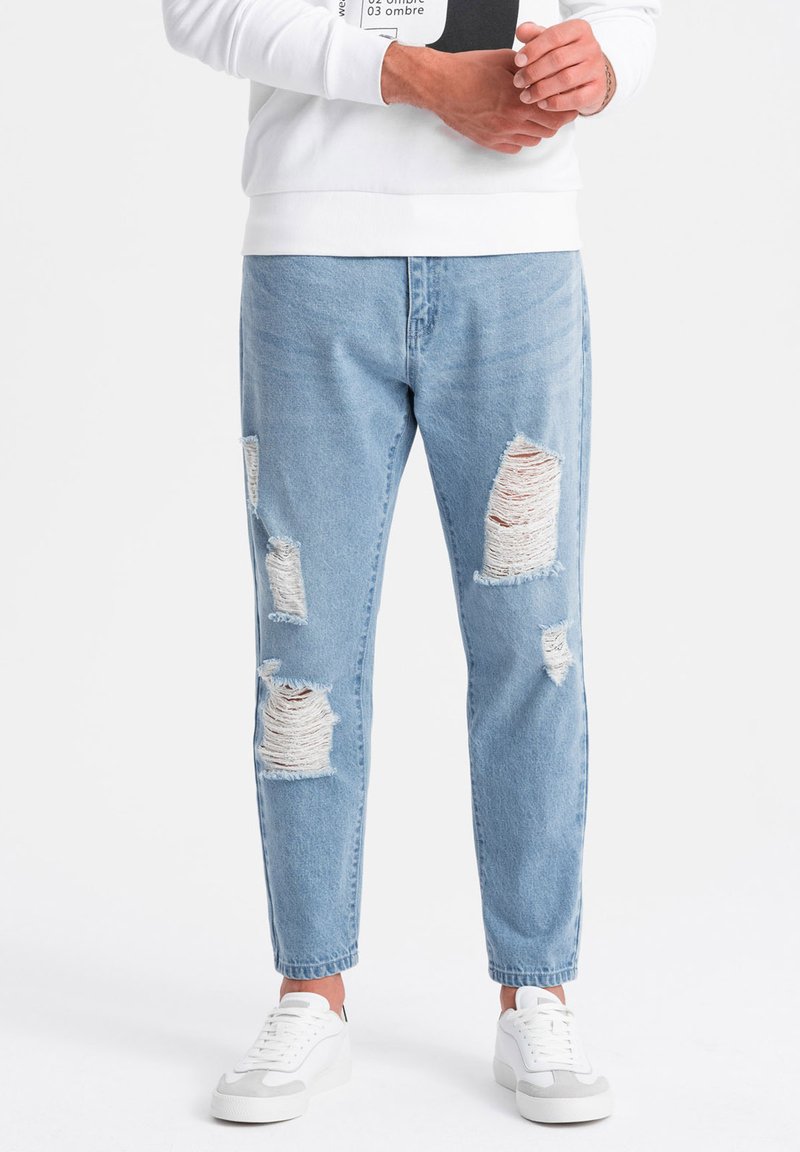Jeans de mezclilla azul claro con rotos, que presentan varios parches grandes desgastados, tobillos doblados y un lavado desgastado, combinados con zapatillas blancas.
