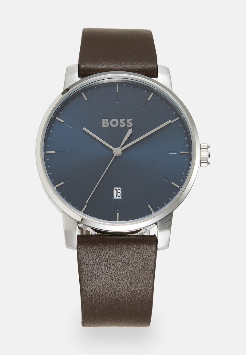 Reloj de pulsera con un dial azul oscuro, caja de tono plateado y correa de cuero marrón. Presenta marcadores de hora minimalistas y una pantalla de fecha a las 6 en punto.