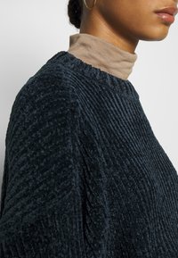 Granatowy sweter o fakturze z pionowym prążkowaniem, z okrągłym dekoltem. Noszony razem z jasnobrązowym golfem pod spodem.