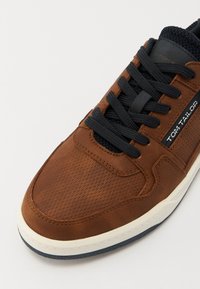 Barna szintetikus felső résszel rendelkező sneaker, textúrázott felülettel, tengerészkék fűzőkkel és fehér középtalppal. Az oldalon a márka logója található.