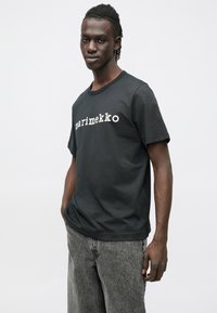 Μαύρο βαμβακερό μπλουζάκι με λευκό λογότυπο "marimekko" στο στήθος. Κοντά μανίκια, λαιμόκοψη γιακά, και άνετη εφαρμογή.