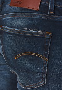 Jeans i mörkblå denim med synlig blekning. Har orangea sömmar och en brun läderetikett på bakre midjan. Detaljerad design av bakfickorna.