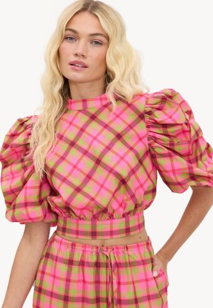 Femme blonde portant un top à manches bouffantes à carreaux roses et verts et un pantalon assorti, posant avec une main dans la poche sur fond blanc.