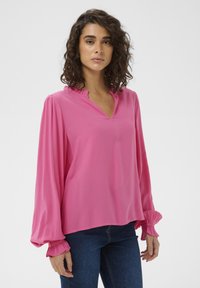 Blouse rose à manches longues avec un col en V et des détails à volants au niveau du col et des poignets. Tissu doux, coupe ample, portée avec un jean bleu.