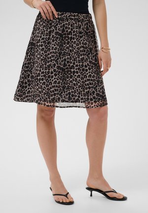 Kvinde iført en knælang nederdel med leopardprint, sort top, guldbøjler, ringe og sorte hælede sandaler på en lys baggrund.