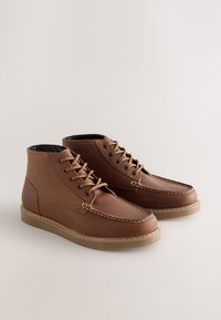 Bottes basses en cuir marron avec coutures contrastées, bout rond, semelle en caoutchouc plate et conception à lacets. Dotées d'une doublure intérieure noire.