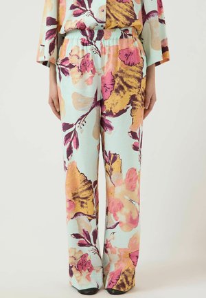 Løse lyseblå bukser med stort pink, lilla og gult blomsterprint, båret med matchende top og sorte sko på en neutral baggrund.