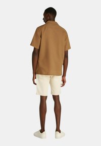 Camicia marrone a maniche corte con una vestibilità rilassata, abbinata a pantaloni corti beige chiaro e sneakers bianche, vista da dietro. Design semplice, tessuto texturizzato.