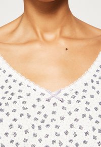 Camiseta de tela blanca con pequeños patrones florales morados, un escote ribeteado con encaje y un lazo de color morado claro en el centro.