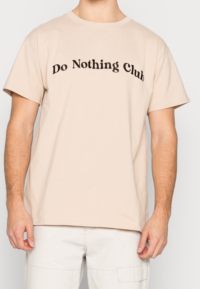 Béžové tričko s krátkým rukávem a černým textem "Do Nothing Club" oblečené osobou se světlou pletí a světlými kalhotami.