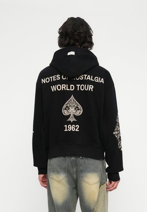 Oseba s temno lasmi do ramen, oblečena v črn kapuzen hoodie z napisom "NOTES OF NOSTALGIA WORLD TOUR 1962" in okrasnim motivom trefla na hrbtu.