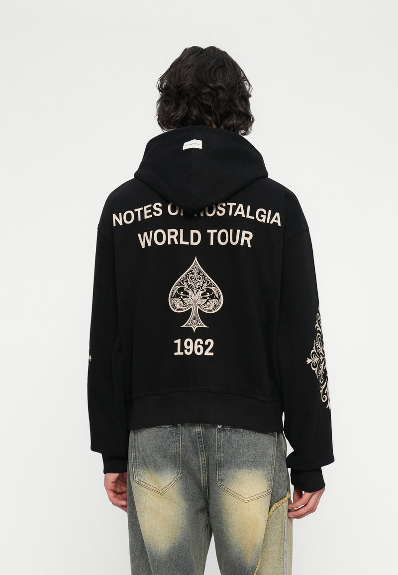 Osoba s tamnom do ramena tamnom kosom koja nosi crni duksericu s natpisom "NOTES OF NOSTALGIA WORLD TOUR 1962" i dekorativnim dizajnom pika na leđima.