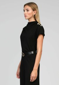 Robe noire à manches courtes, col haut et quincaillerie décorative sur l'épaule. Taille ajustée accentuée par une ceinture noire avec une boucle en or.