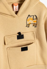 Felpa con cappuccio beige con un'illustrazione di un cane arancione e marrone, dotata di una tasca frontale con dettagli in velcro nero. Materiale tessile morbido.