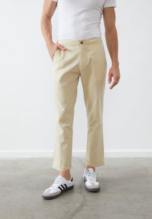Mann trägt beige gerade geschnittene Hose mit umgeschlagenen Säumen, weißes T-Shirt und weiße Sneakers mit schwarzen Streifen, steht auf grauem Boden.