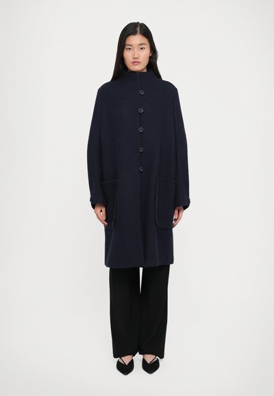 Donna in piedi con un cappotto blu navy con bottoni e grandi tasche frontali, pantaloni neri a gamba larga e scarpe nere a punta su uno sfondo bianco.
